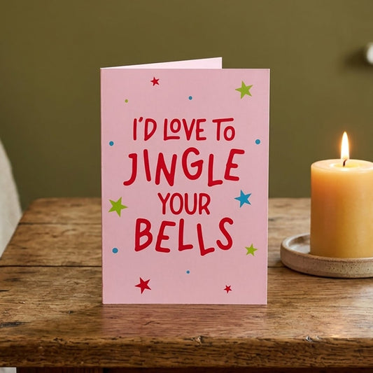 A pink greeting card with the text 'I'D LOVE TO JINGLE YOUR BELLS' in red, decorated with small multicolored stars, standing on a rustic wooden table next to a lit yellow candle.