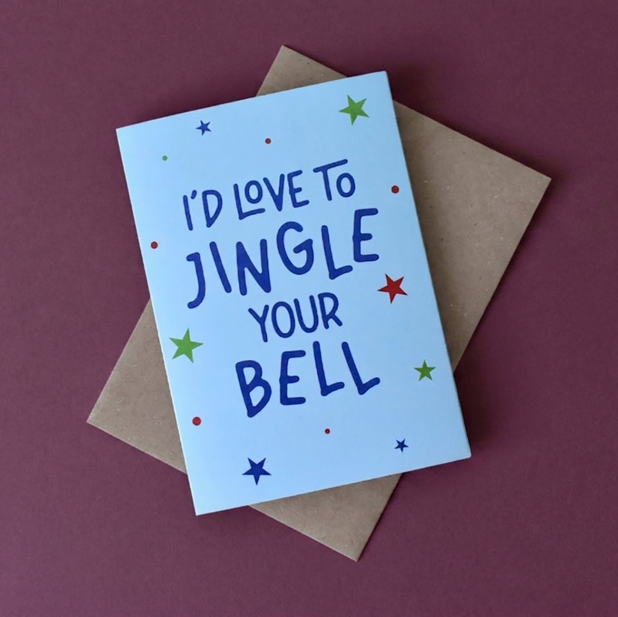 A light blue greeting card with the phrase 'I'D LOVE TO JINGLE YOUR BELL' in dark blue lettering, decorated with small colorful stars and dots, resting on a brown kraft envelope against a solid dark plum background.