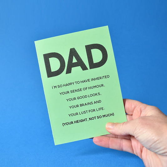 A humorous greeting card addressed to 'DAD' with printed text listing positive inherited qualities, placed on a table with a plant in the background.