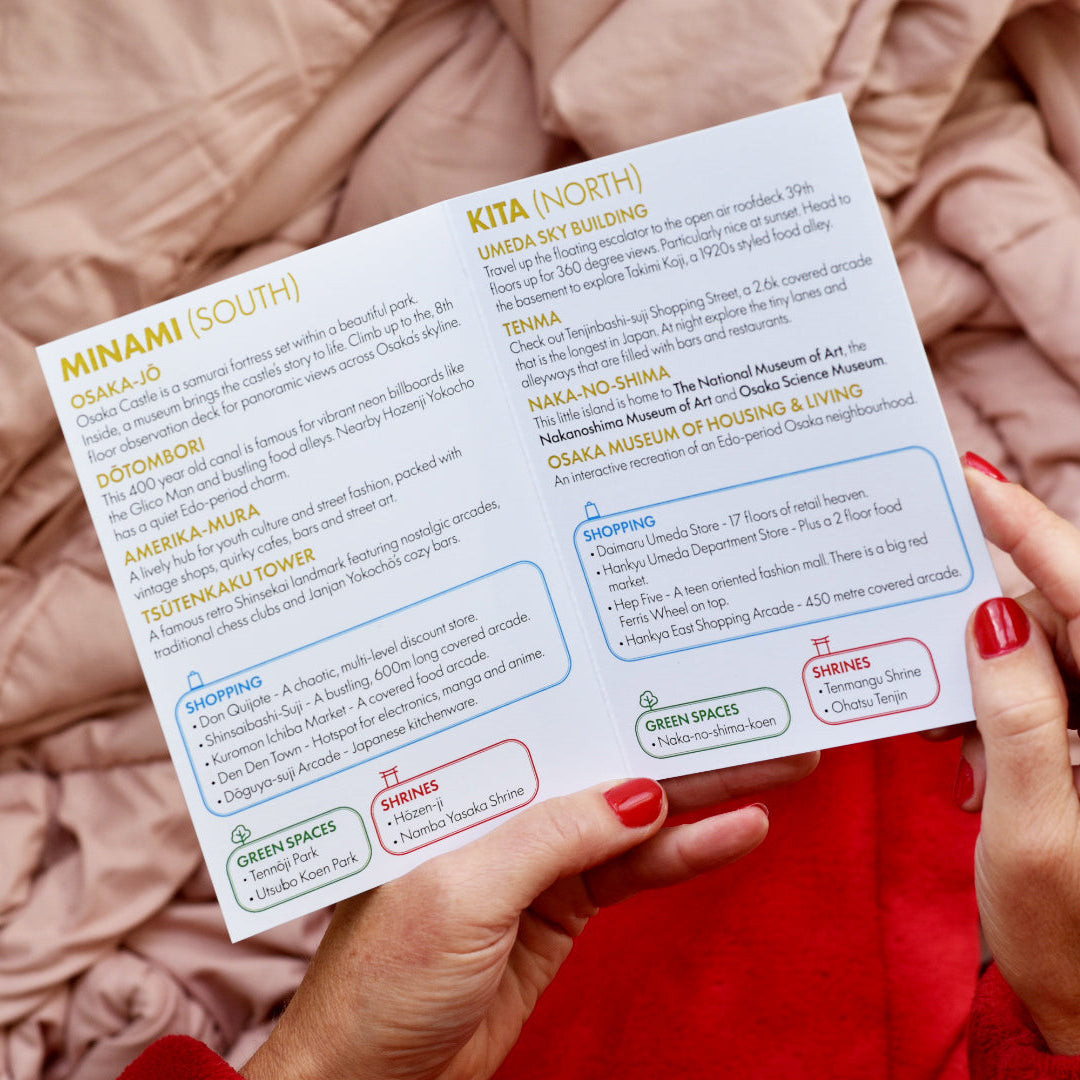 A person holds an open printed booklet showing neighbourhood information for Minami and Kita, with hands and a pink bedspread visible in the background.