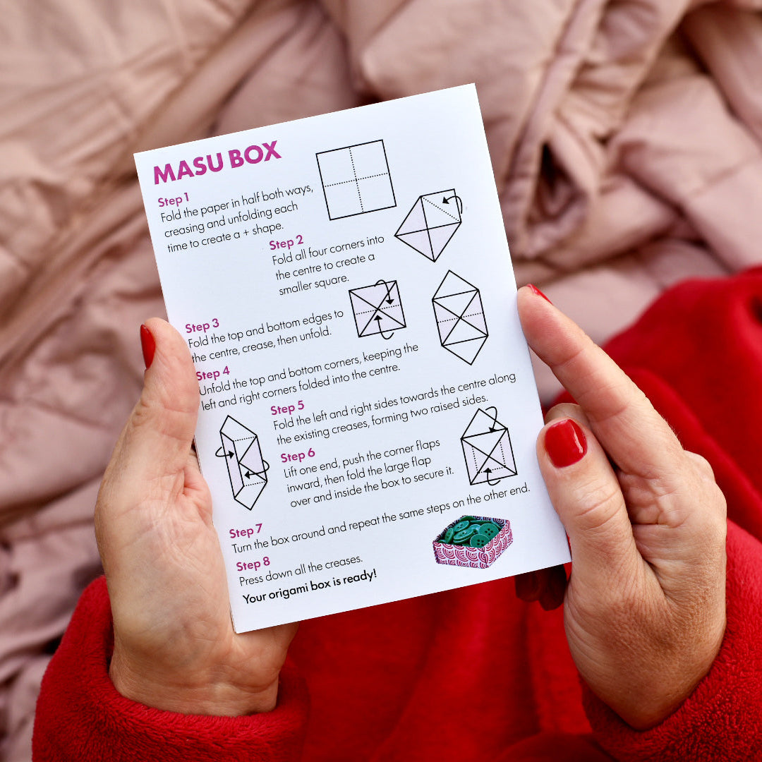 A person holding a printed folding instruction sheet for a MASU BOX origami box, with step-by-step diagrams and text on a white card. The person is wearing a red sweater and holding the card over a pink blanket background.