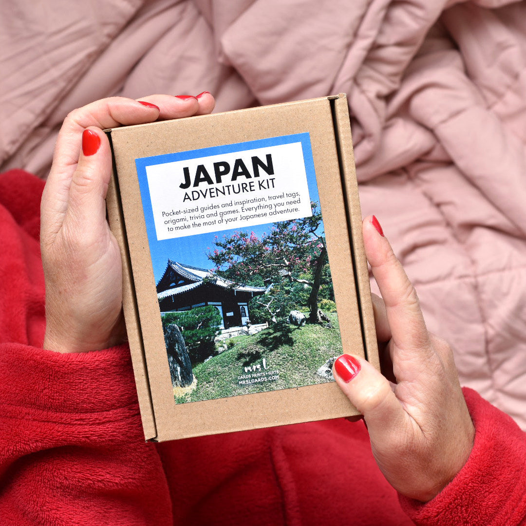 A person holding a small brown cardboard box labeled 'JAPAN ADVENTURE KIT' with a printed cover photo, against a soft pink blanket background.