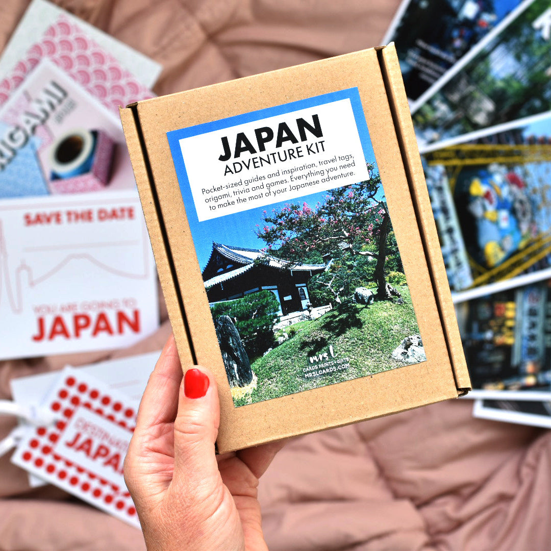 A hand with red nail polish holds a brown cardboard box labeled 'JAPAN ADVENTURE KIT' in front of a spread of Japan-themed cards and travel prints on a pink quilted blanket.