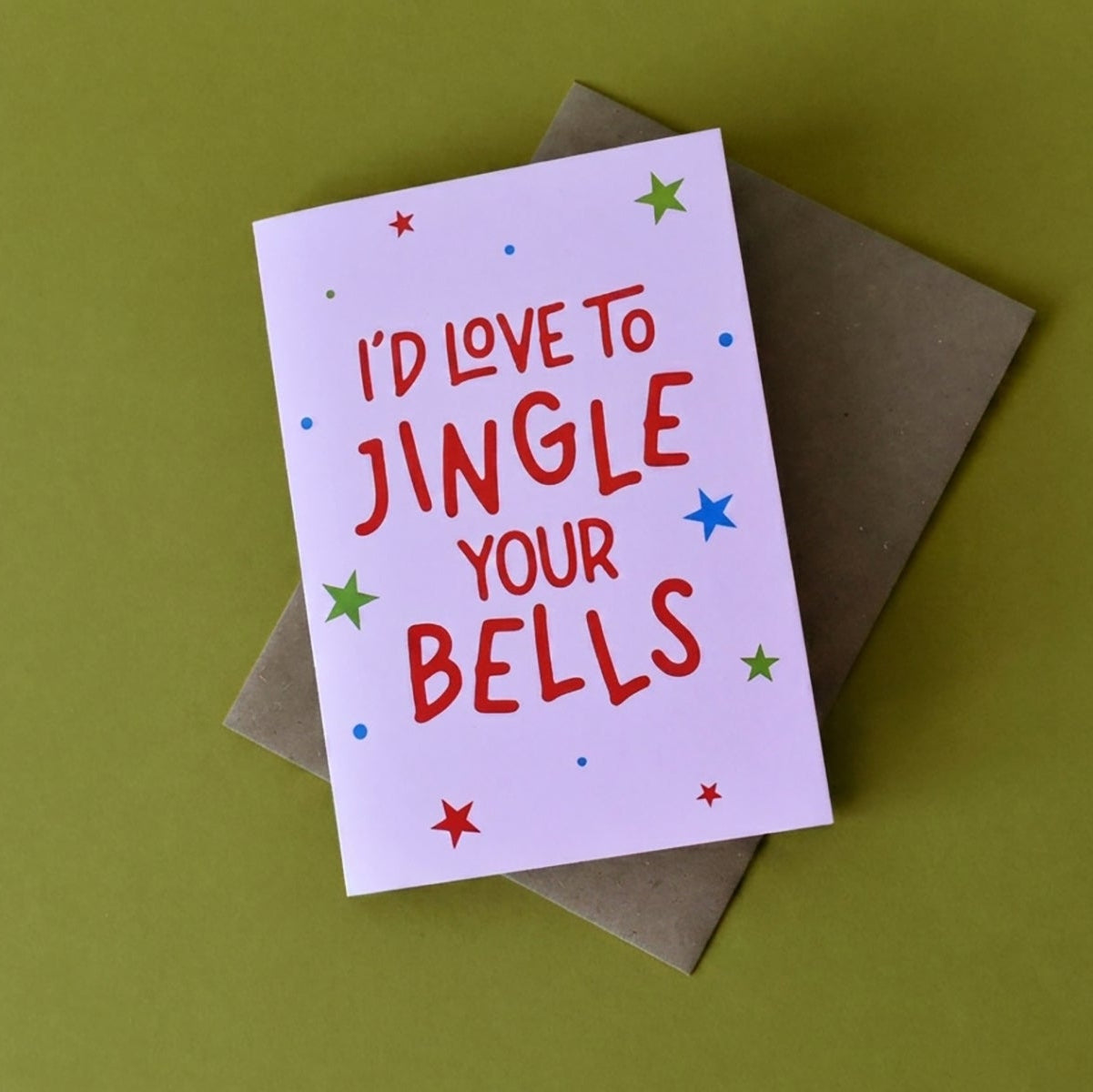 A square greeting card with the phrase 'I'D LOVE TO JINGLE YOUR BELLS' in red lettering, decorated with small multicolored stars and dots on a pale pink background. The card is placed on top of a brown envelope against an olive green background.