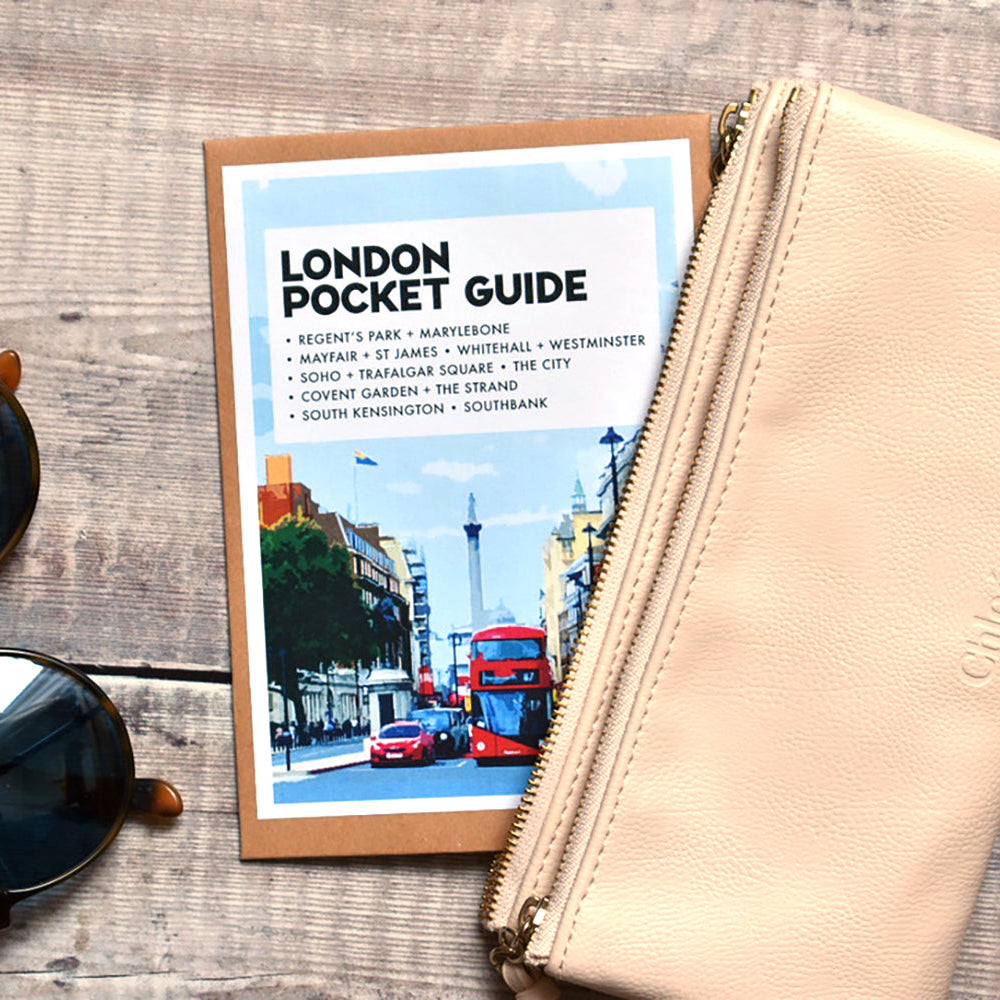 A beige leather zip pouch with an embossed logo, placed next to a 'London Pocket Guide' booklet and a pair of sunglasses on a rustic wooden background.