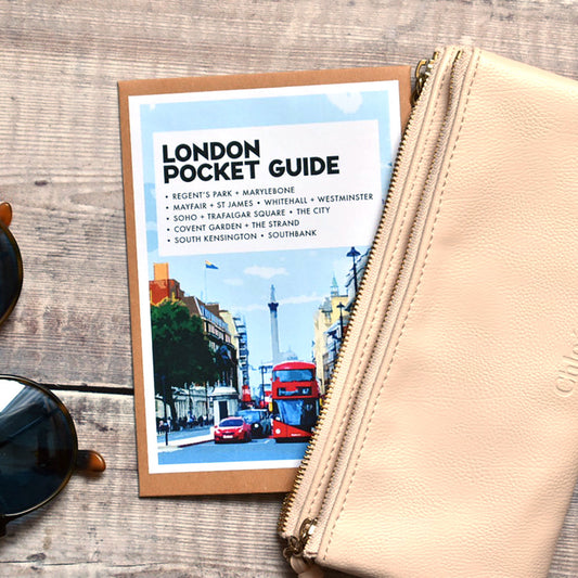 A beige leather zip pouch with an embossed logo, placed next to a 'London Pocket Guide' booklet and a pair of sunglasses on a rustic wooden background.