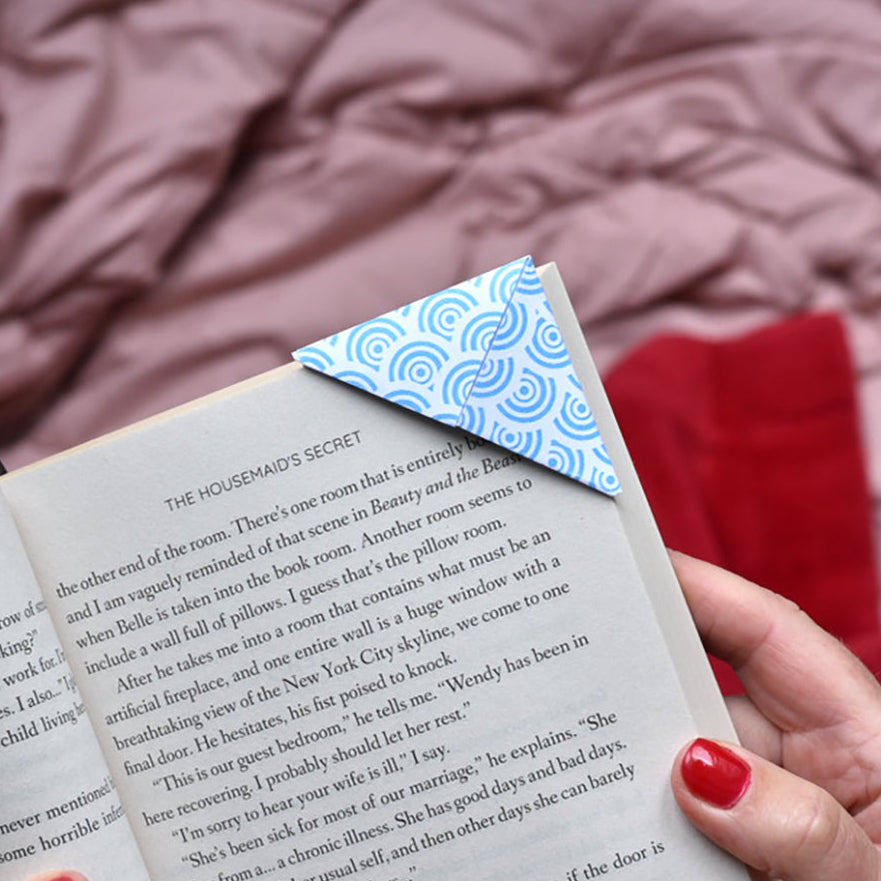 A close-up of hands holding an open book with a blue and white patterned origami bookmark attached to the page, set against a soft pink blanket background with a red fabric accent.