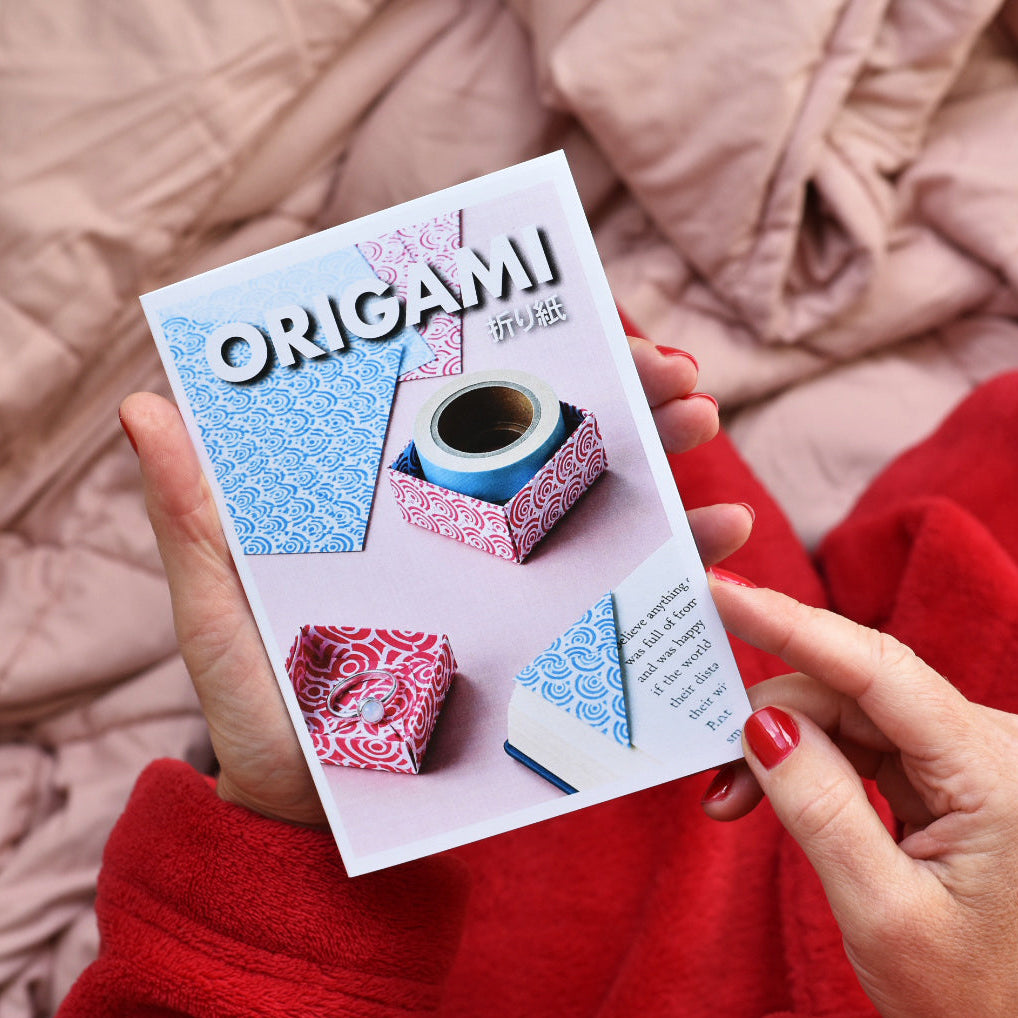 A person holding a Japanese origami booklet titled 'ORIGAMI 初級' with both hands, set against a cozy background of a dusty pink quilted blanket and a red fleece blanket.