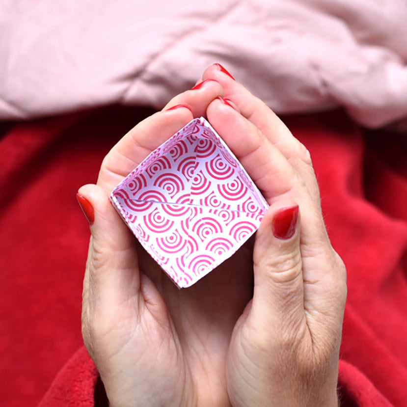 A close-up of a person's hands holding a small pink patterned origami Masu box, with a red blanket and a light pink quilted blanket in the background.