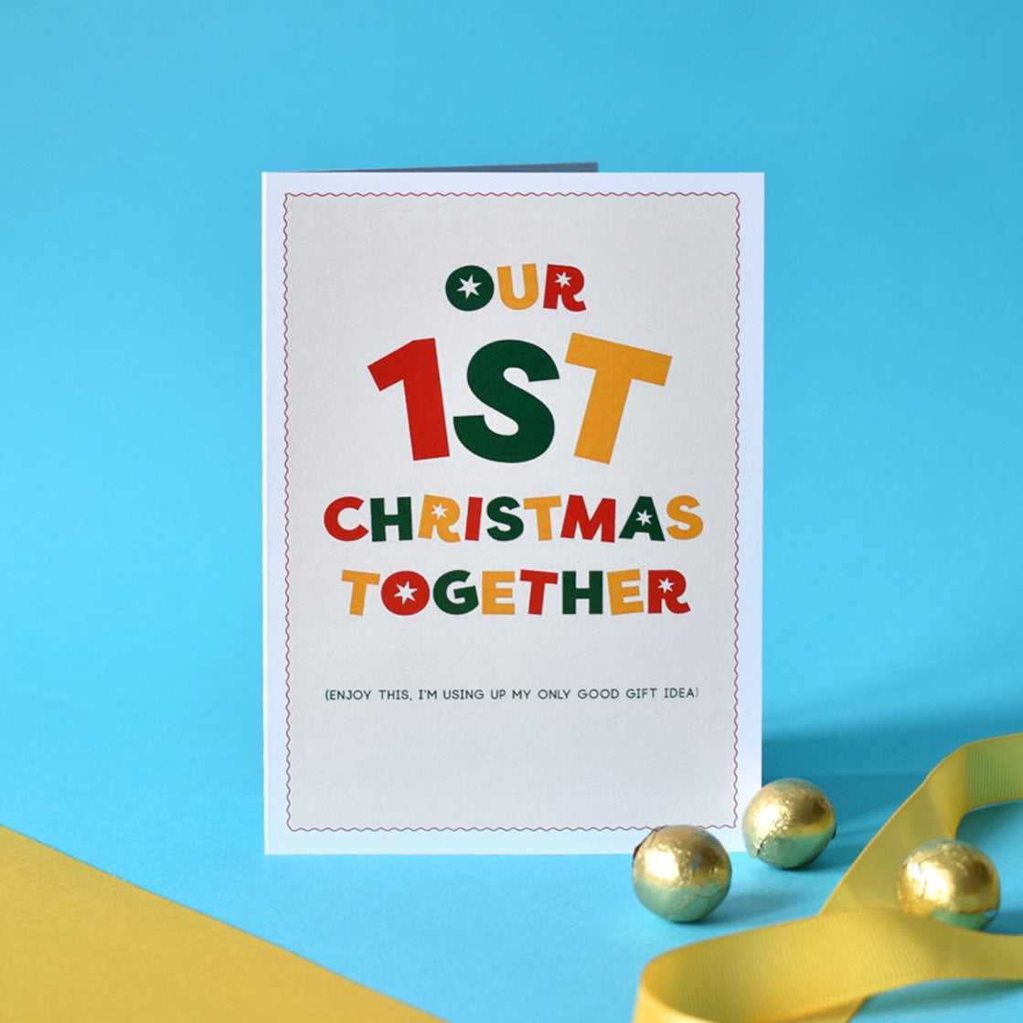 A white greeting card with colorful text reading 'OUR 1ST CHRISTMAS TOGETHER' stands on a bright blue background with gold foil balls and yellow ribbon as props.