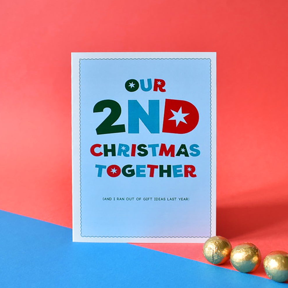 A square greeting card with colorful text reading 'OUR 2ND CHRISTMAS TOGETHER' stands on a blue and red background with three gold ornaments near the bottom right.