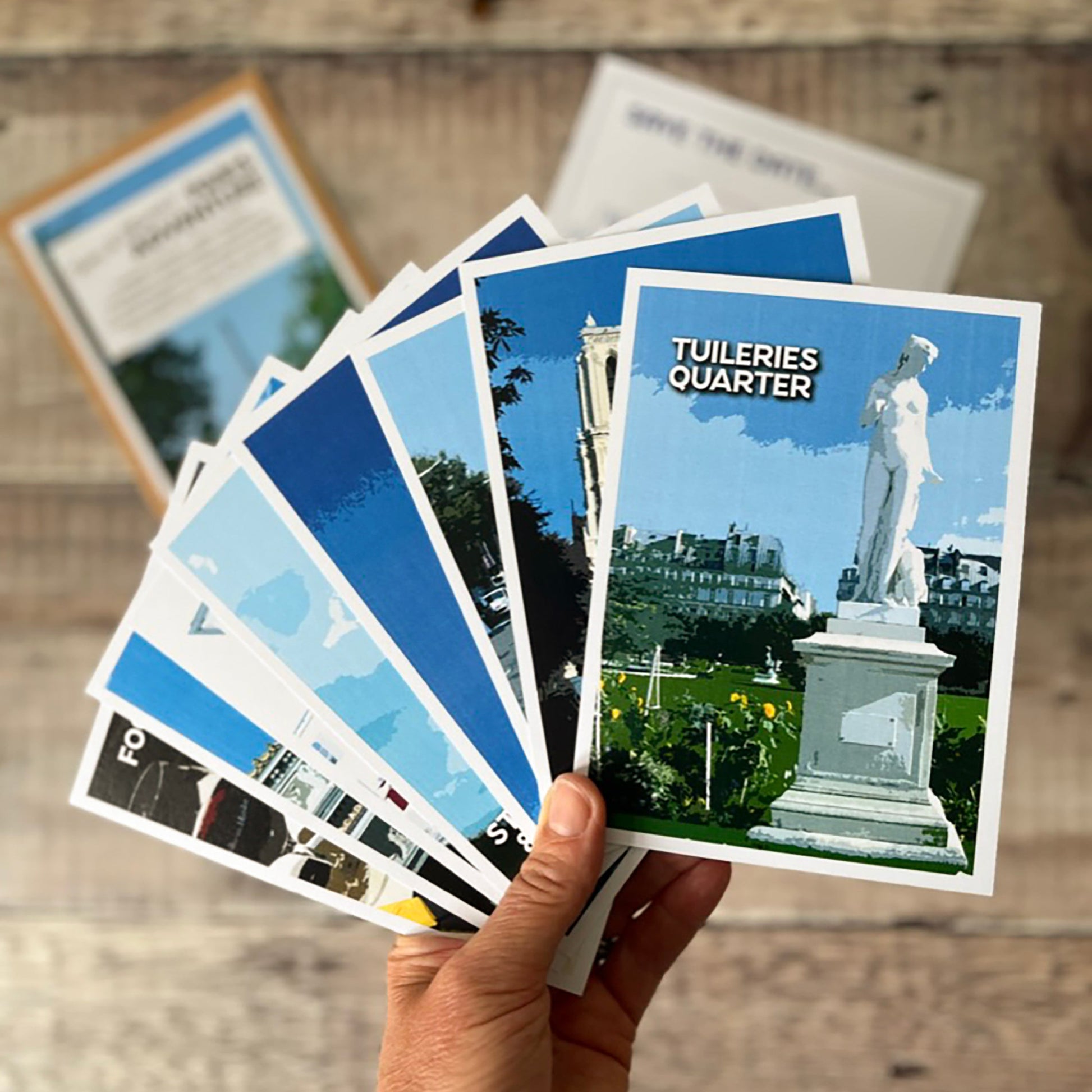 A hand holds a fanned-out set of postcards showing scenic views of the various areas in Paris, with a wooden surface and sunglasses in the background.