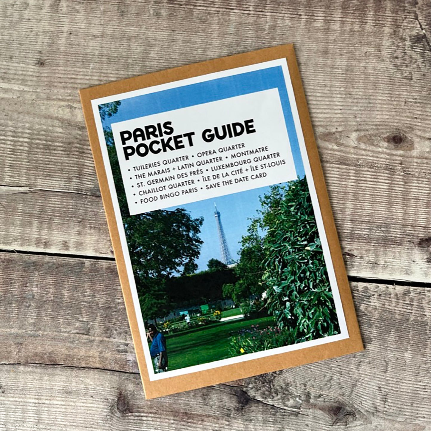 A Paris Pocket Guide postcard with travel cards placed on a rustic wooden surface. The card has a tan border, a blue header, and a photo of a park scene with the Eiffel Tower visible in the background.