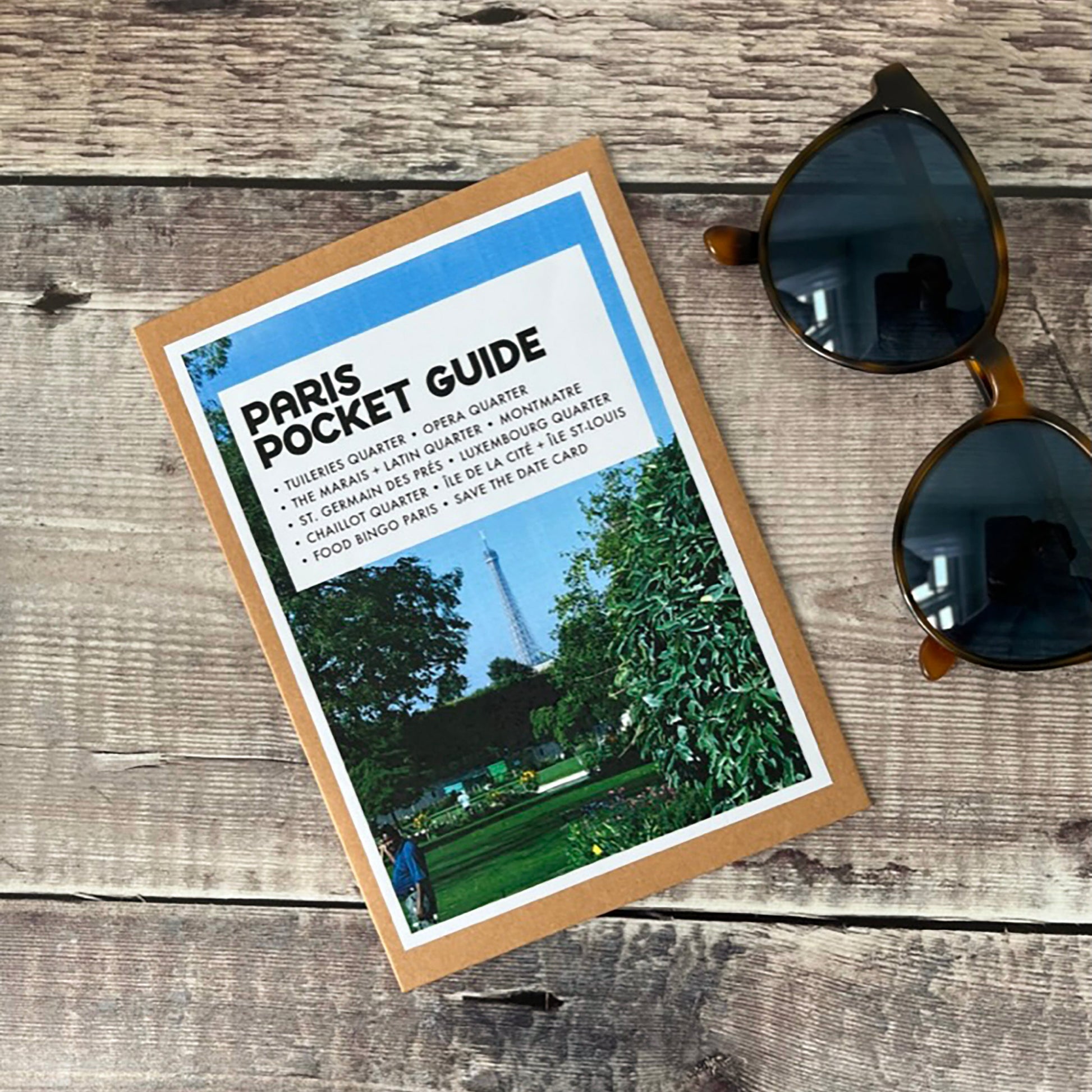 A Paris Pocket Guide booklet lies on a rustic wood plank surface next to a pair of dark sunglasses. The booklet is made from card and has a cover showing a green park scene with blue sky and the Eiffel Tower in the background. A wonderful gift for someone visiting Paris.