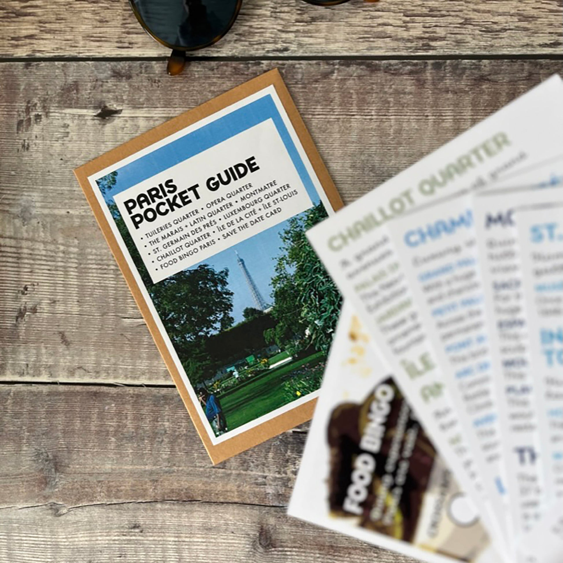 A Paris Pocket Guide booklet lies on a rustic wood surface next to a pair of sunglasses, with a hand holding several printed cards in the foreground labeled 'Chaillot Quarter'.