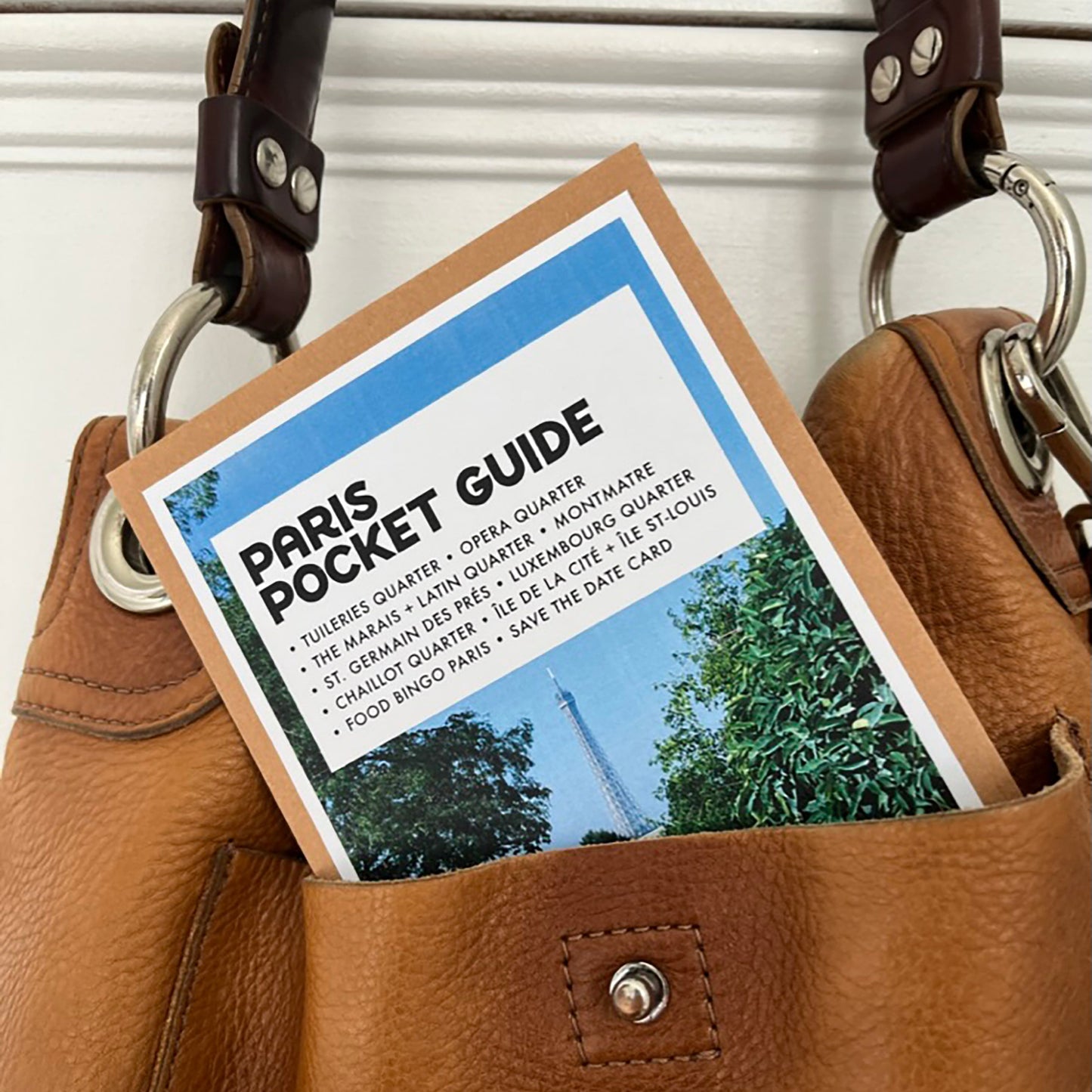 A tan leather shoulder bag with dark brown leather straps and silver-tone hardware, shown holding a Paris Pocket Guide card in its front pocket against a white paneled background.