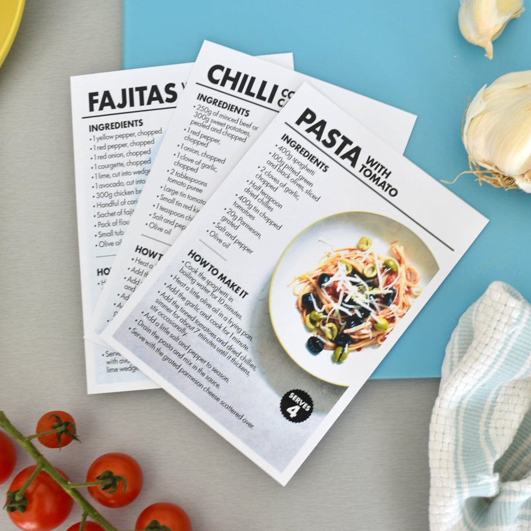 Three printed recipe cards for Fajitas, Chilli Pasta, and Pasta Medalla are arranged on a light gray surface with a bright blue background. The cards are styled with a yellow spoon, cherry tomatoes, garlic, and a striped cloth.