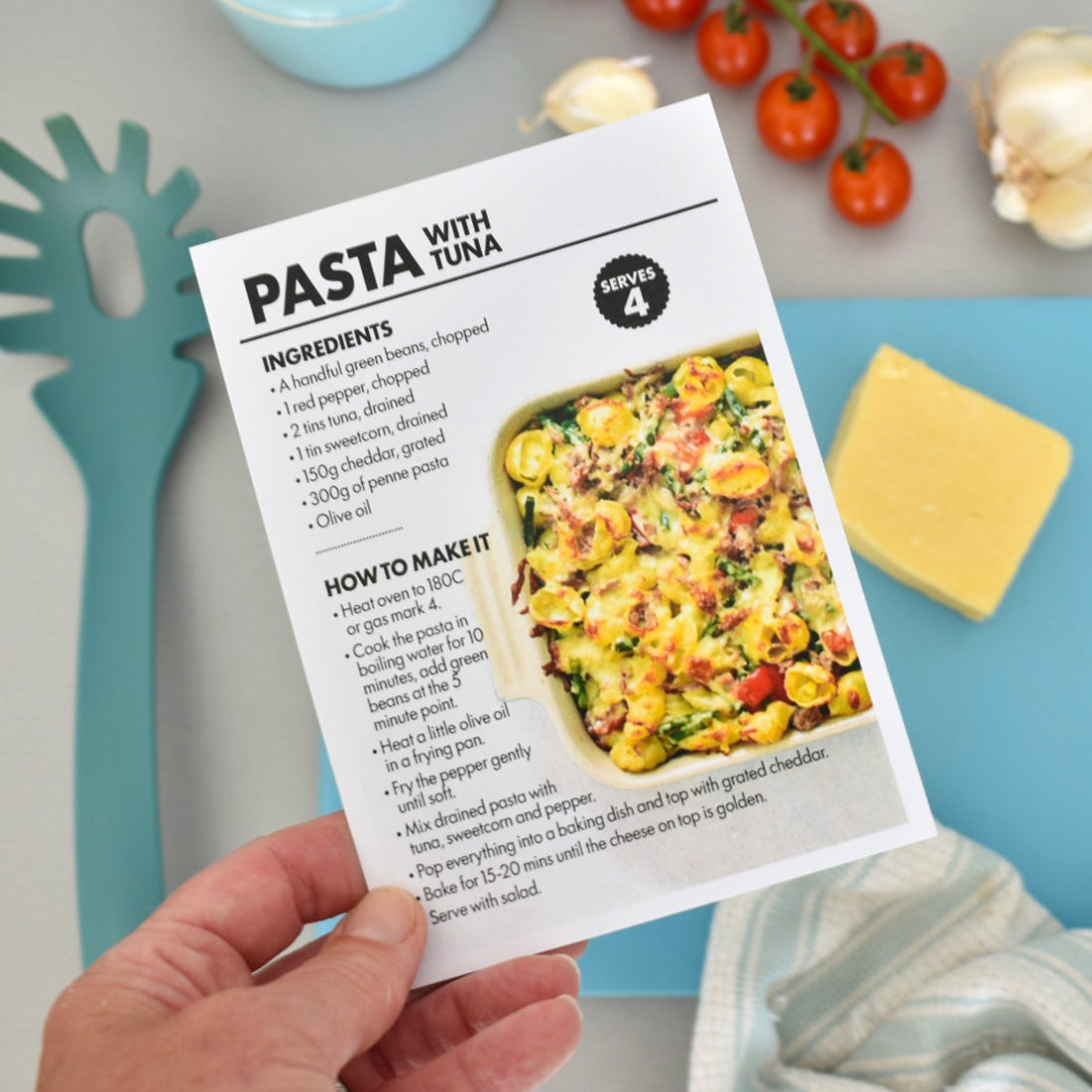 A hand holds a printed pasta recipe card titled 'PASTA WITH TUNA' over a styled kitchen surface with a blue cutting board, garlic, tomatoes, and a striped cloth.