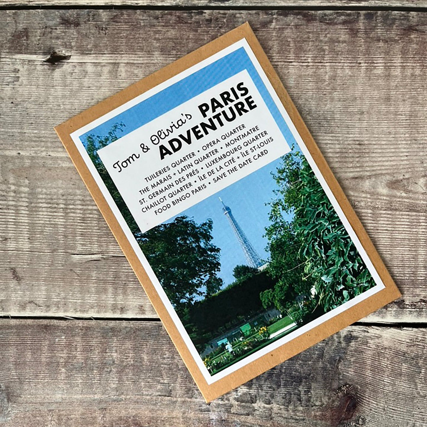 A square kraft paper card with a photo of Paris and the text 'Tom & Olivia's PARIS ADVENTURE' lies on a rustic wooden surface.