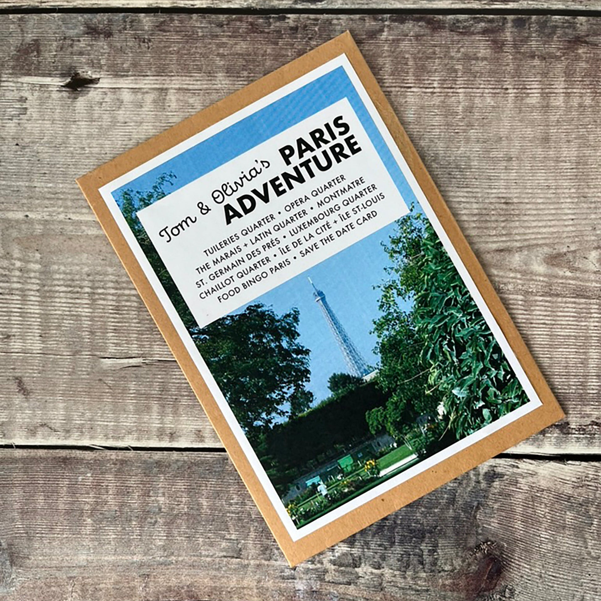 A square kraft paper card with a photo of Paris and the text 'Tom & Olivia's PARIS ADVENTURE' lies on a rustic wooden surface.