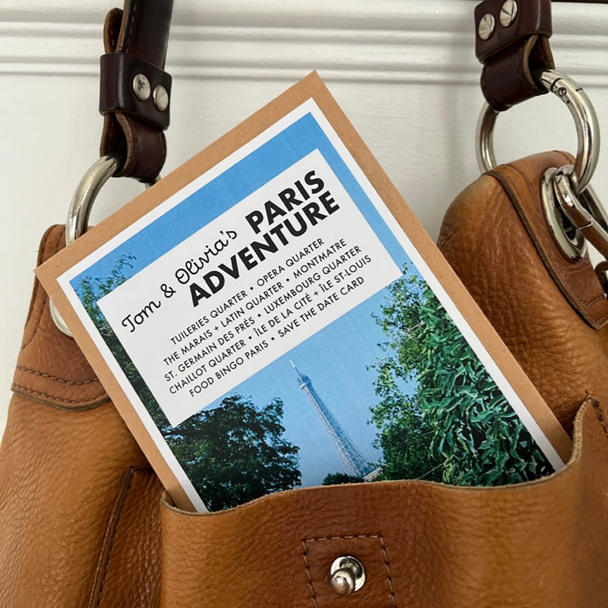 A tan leather handbag with dark brown leather straps and silver-tone hardware, shown holding a printed Paris adventure card or postcard in its front pocket against a white paneled background.