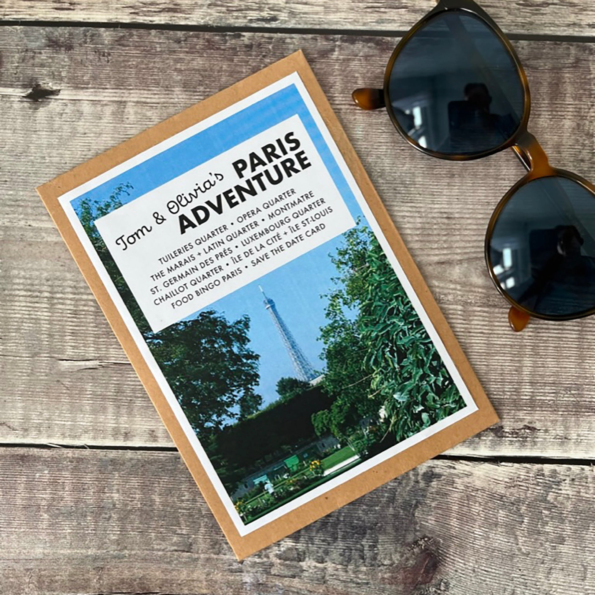 A flat-lay photograph of a kraft card wallet featuring a Paris adventure travel guides  styled on a rustic wooden background next to a pair of dark sunglasses.
