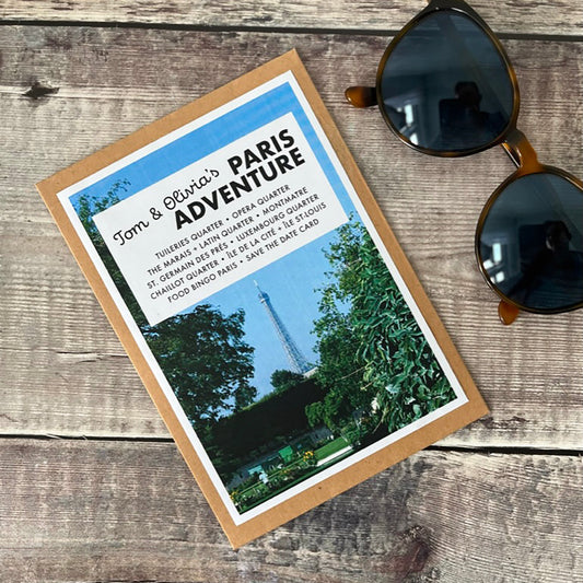 A flat-lay photograph of a kraft card wallet featuring a Paris adventure travel guides  styled on a rustic wooden background next to a pair of dark sunglasses.
