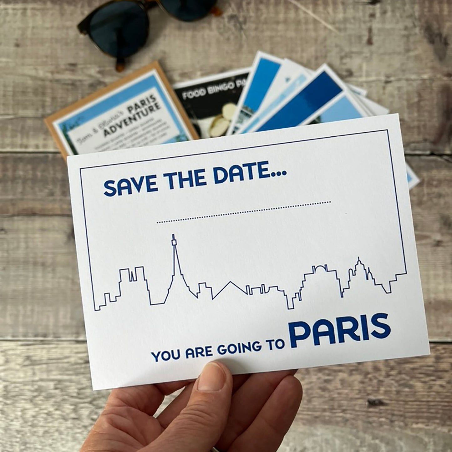 A hand holds a white 'Save the Date' Paris-themed card with a blue skyline outline and text reading 'SAVE THE DATE, You Are Going to Paris.
