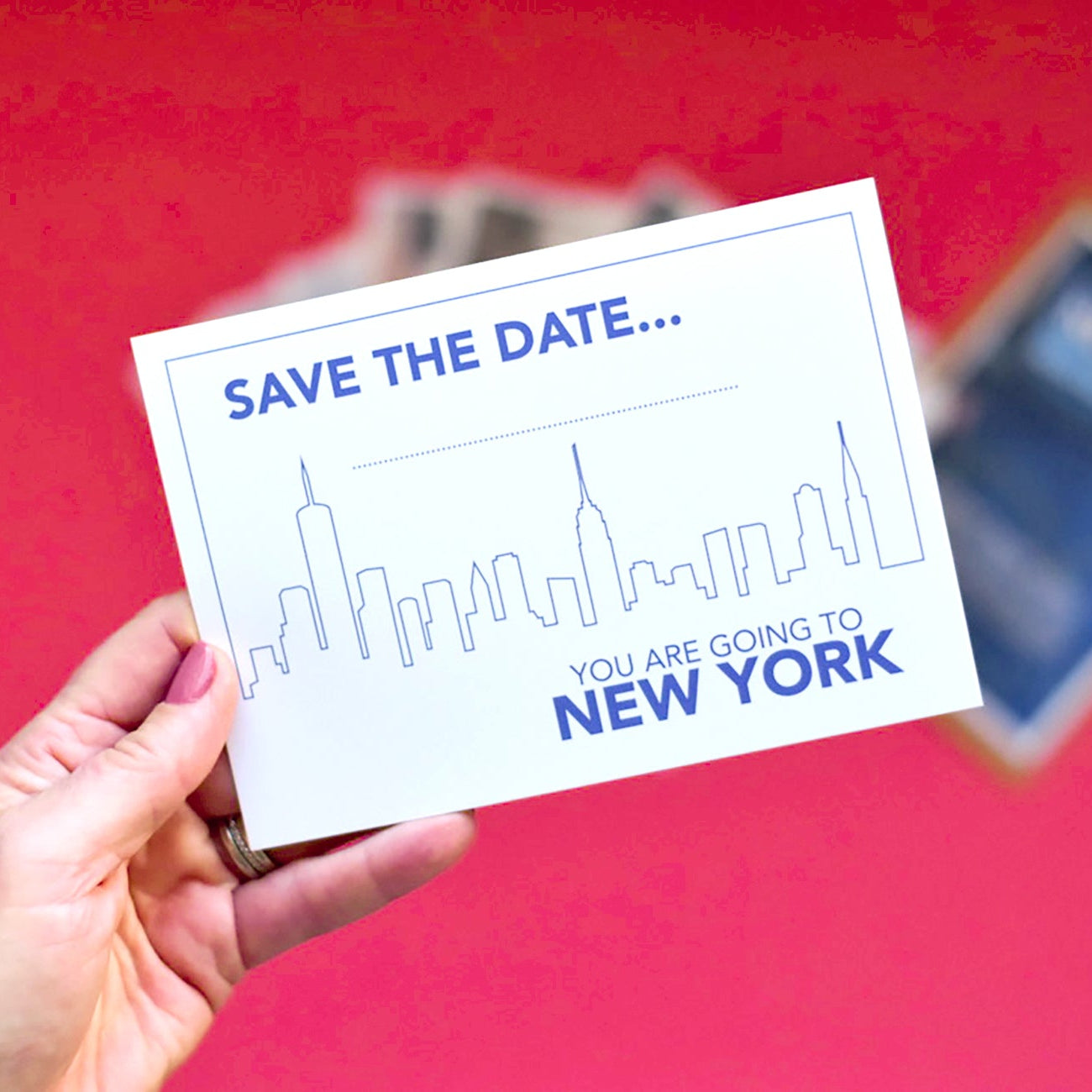 Hand holding a 'Save the Date' card with New York City skyline design against a red background