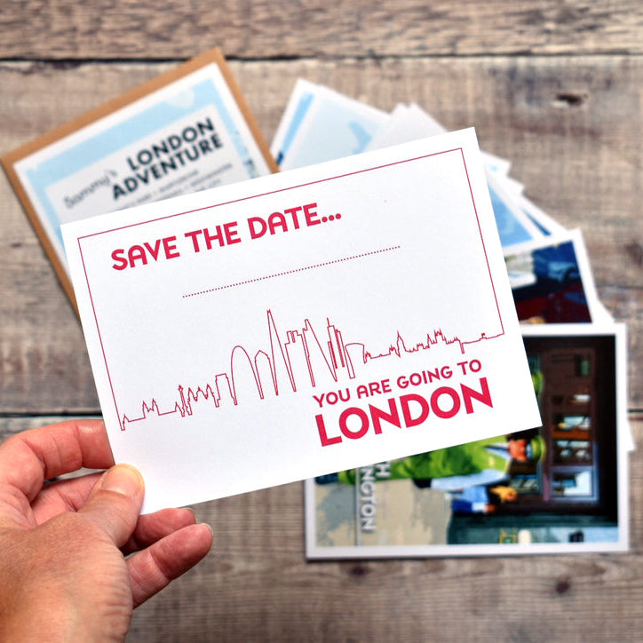 A hand holds a white postcard with red text reading 'SAVE THE DATE, YOU ARE GOING TO LONDON.