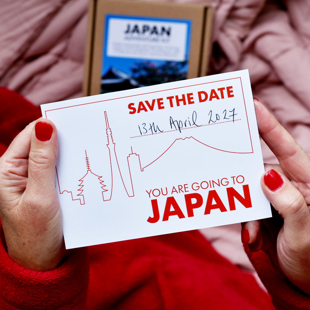 A person holding a white 'Save the Date' card with red text and a red line-art skyline, with a brown box labeled 'You are going to JAPAN' in the background against pink bedding.