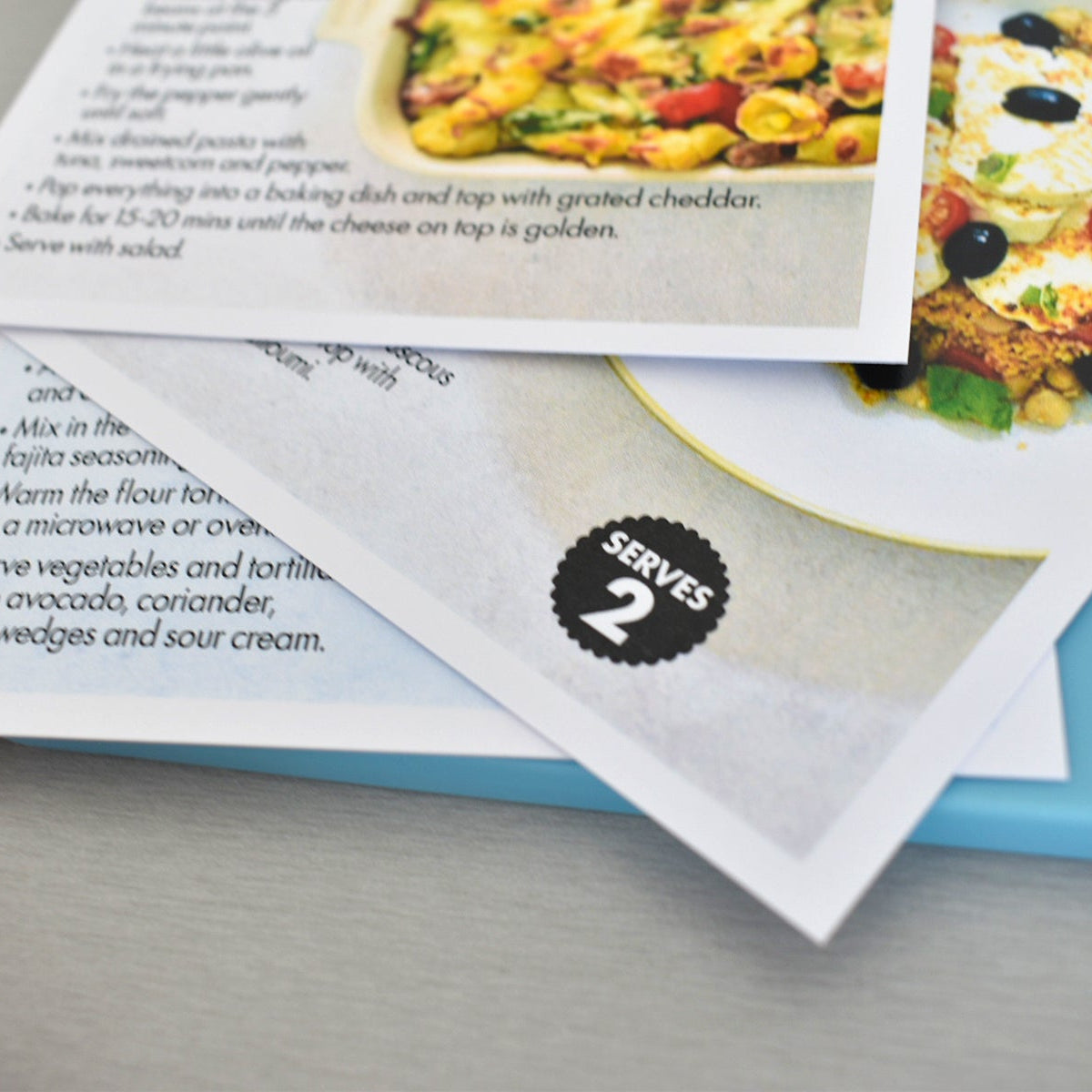 A close-up of printed recipe cards or instruction sheets layered on a light blue folder, with food photography and text partially visible.