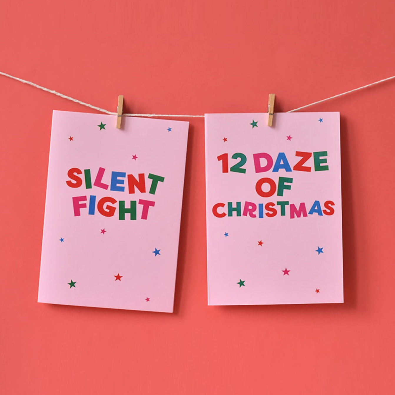 Two pink cards with Christmas-themed text hanging on a string against a red background. The wording reads "Silent Fight" and "12 Daze of Christmas". 