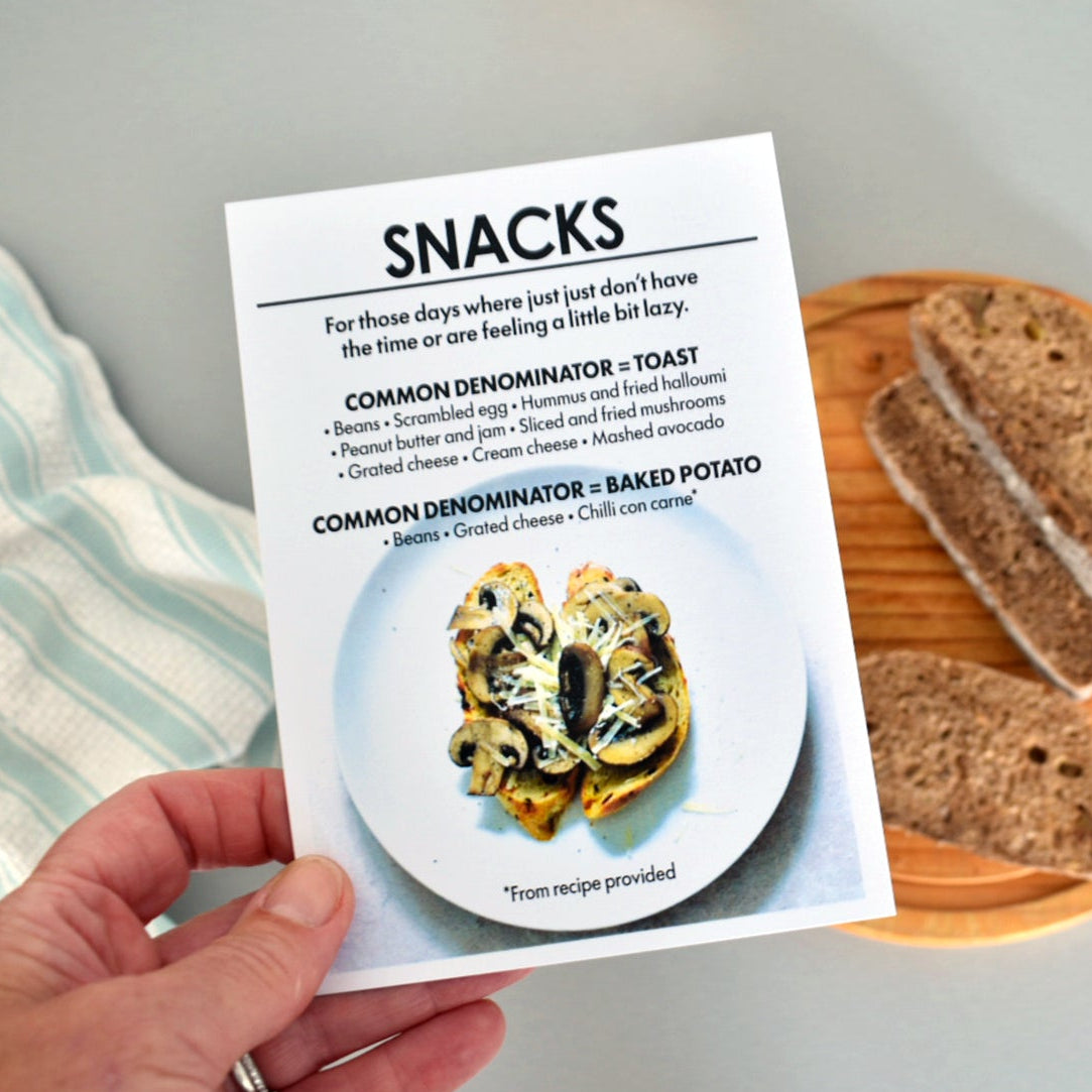 A hand holds a printed snack recipe card titled 'SNACKS' with a photo of stuffed tortilla chips on a plate. In the background, a wooden board holds slices of bread on a light gray surface.