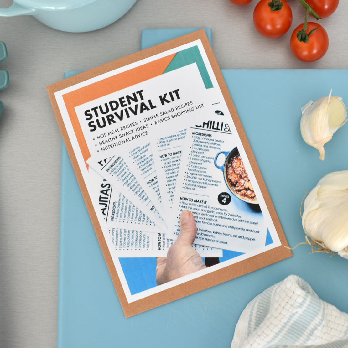 A hand holding a printed 'Student Survival Kit' booklet over a light blue cutting board, surrounded by kitchen props including tomatoes, garlic, a pasta server, and a towel.