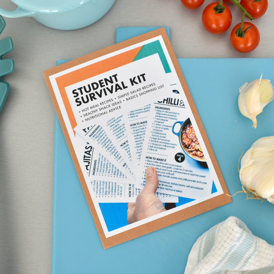 A hand holding an open booklet titled 'Student Survival Kit' on a blue cutting board, surrounded by kitchen props including a teal pasta server, tomatoes, garlic, and a striped cloth on a gray surface.