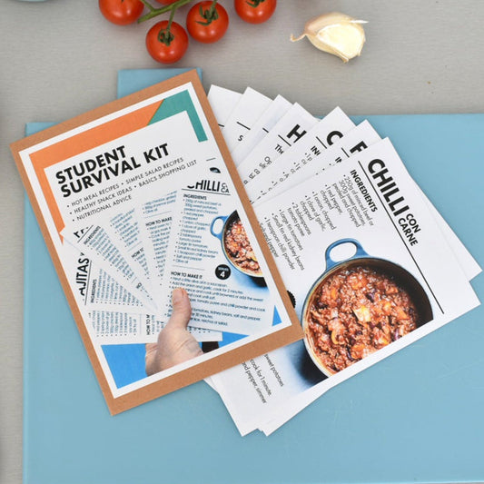 A styled flat-lay of a Student Survival Kit booklet and printed recipe cards on a light blue folder, surrounded by kitchen props including tomatoes, garlic, a teapot, a spatula, and a striped cloth on a gray surface.