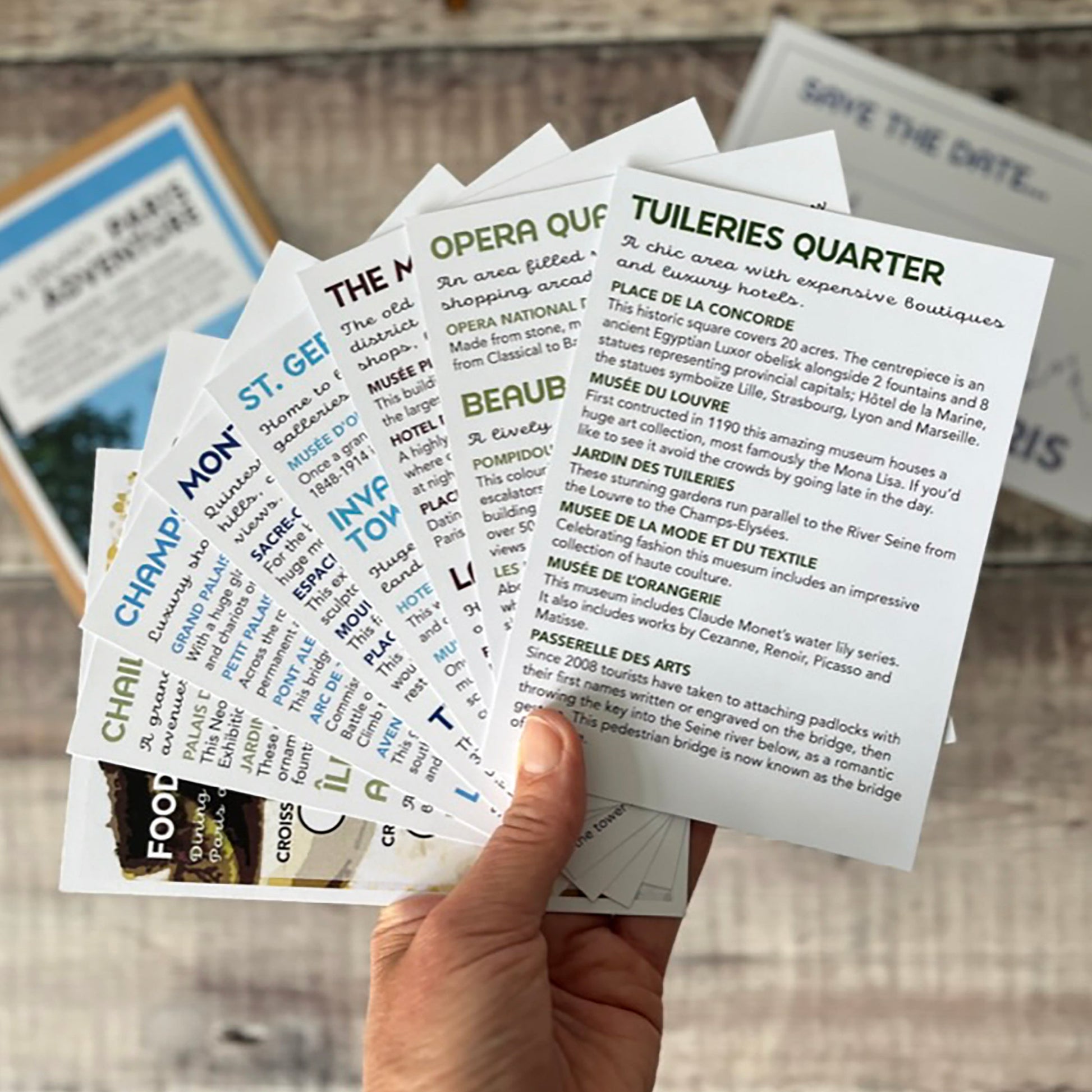 A hand holds a fanned-out set of printed Paris cards for places such as Opera Quarter, Tuileries Quarter and St. Germain, photographed on a rustic wood surface with sunglasses and other printed items in the background.