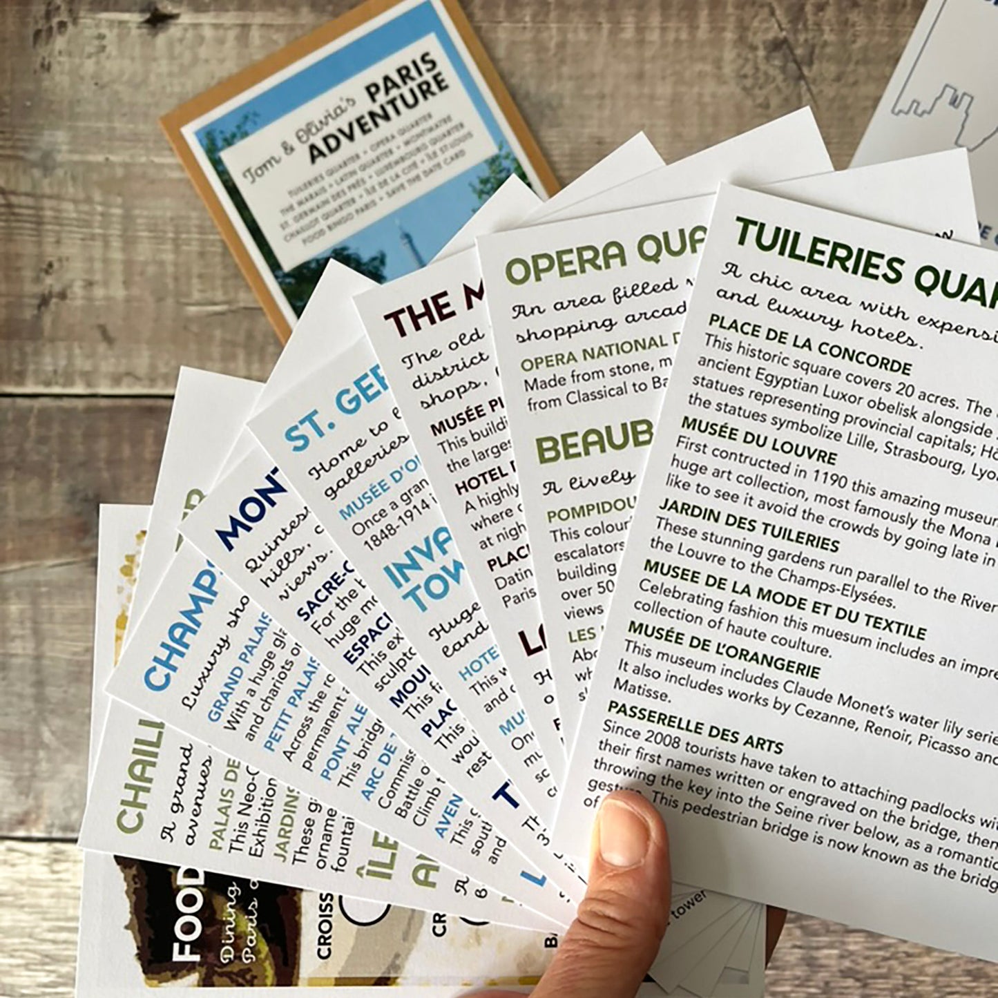 A hand holds a fanned-out set of Paris-themed travel guide cards on a rustic wooden table. The cards feature colourful headers for attractions, food, and landmarks, with detailed descriptions underneath.