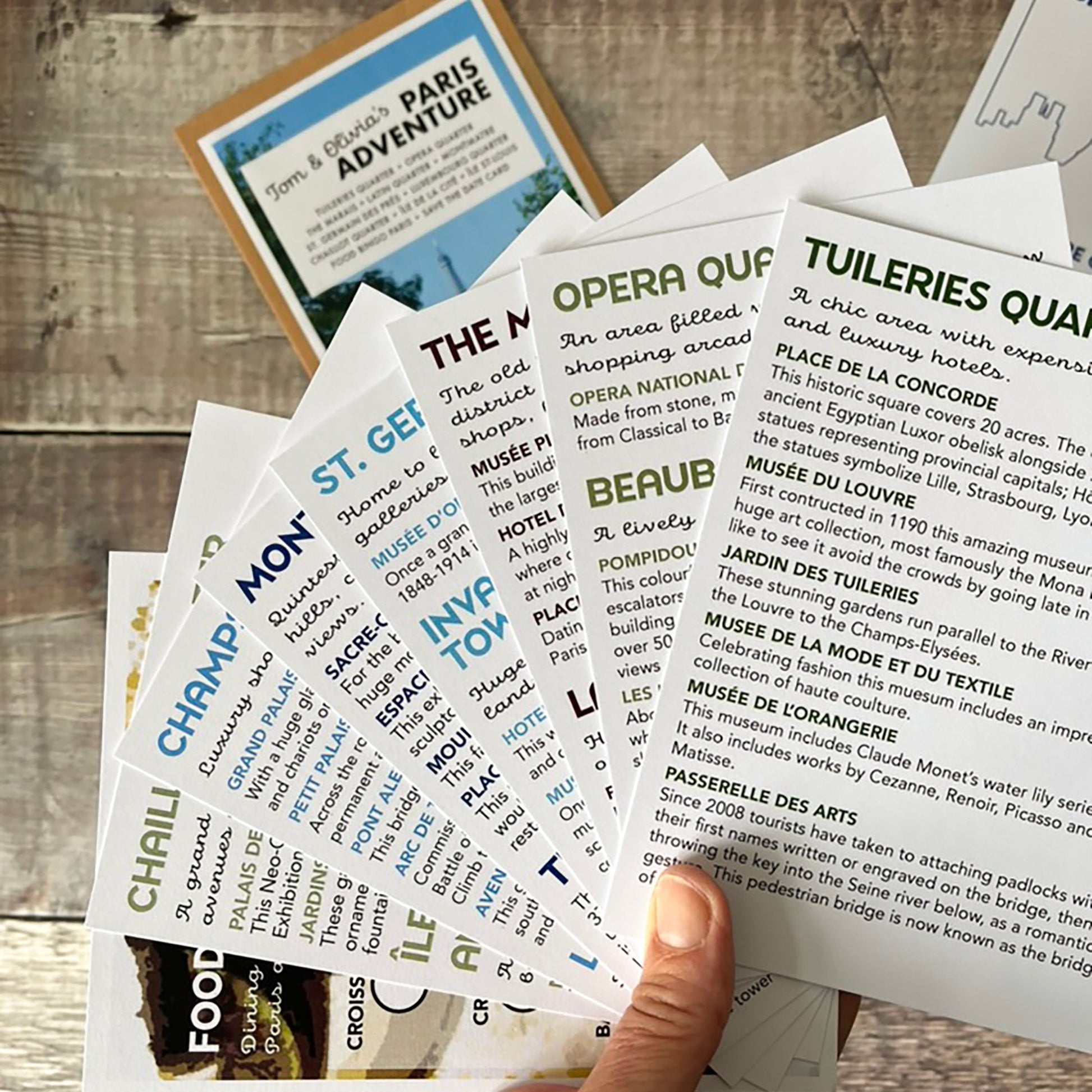A hand holds a fanned-out set of Paris-themed travel guide cards on a rustic wooden table. The cards feature colourful headers for attractions, food, and landmarks, with detailed descriptions underneath.