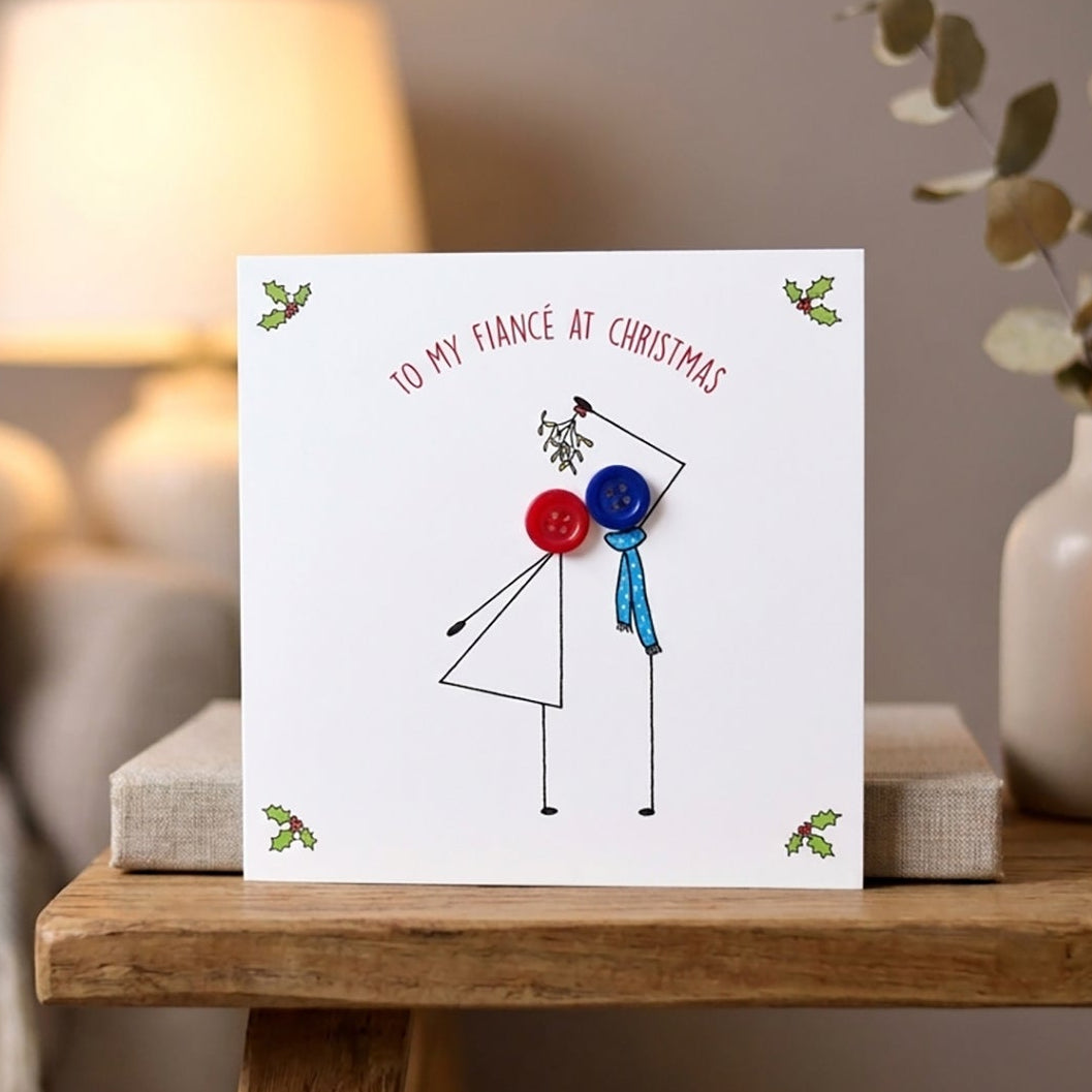 A white Christmas greeting card with a minimalist stick-figure couple and the text 'TO MY FIANCÉ AT CHRISTMAS' in red, styled on a wooden table in a cozy living room setting.