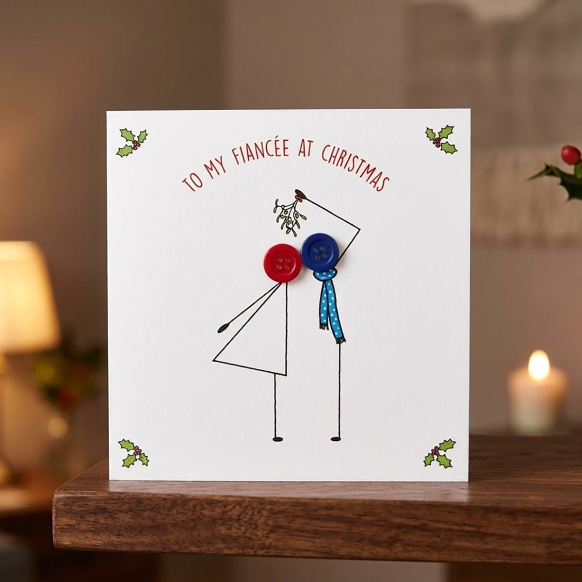 A square white greeting card with a minimalist stick-figure style illustration of a couple and the text 'TO MY FIANCÉE AT CHRISTMAS' at the top, displayed on a wooden table in a warmly lit indoor setting with a lamp, candle, and green vase in the background.