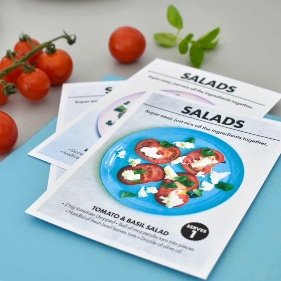 Printed salad recipe cards fanned out on a light blue cutting board, with cherry tomatoes and a small green herb sprig nearby on a gray tabletop.