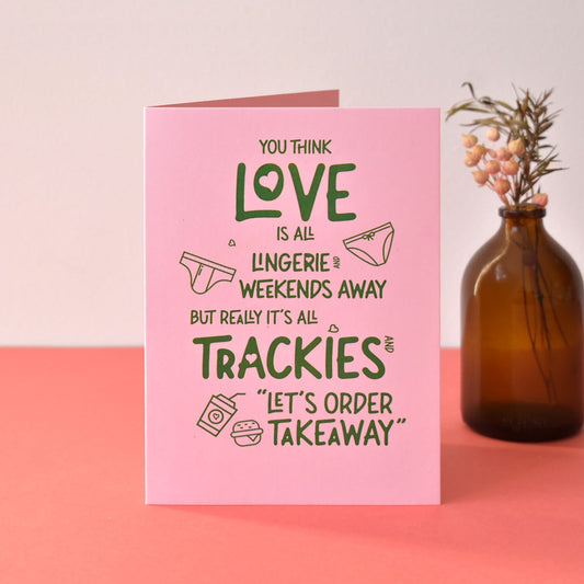 Pink Valentines Card with the text "You think love is all lingerie and weekends away but really its all trackies and let's order takeaway" in green alongside illustrations of underwear and takeaway boxes.