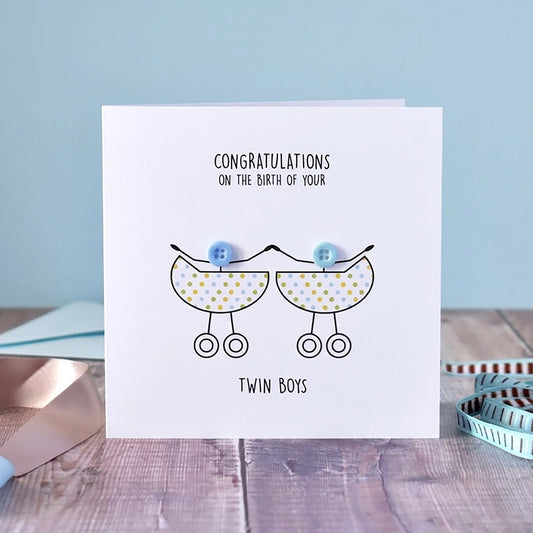 A white square greeting card standing on a wooden surface, printed with the message 'CONGRATULATIONS ON THE BIRTH OF YOUR Twin Boys' and a simple line drawing of two baby prams with blue balloons. The card is styled with a pale blue background, ribbons, and a measuring tape.