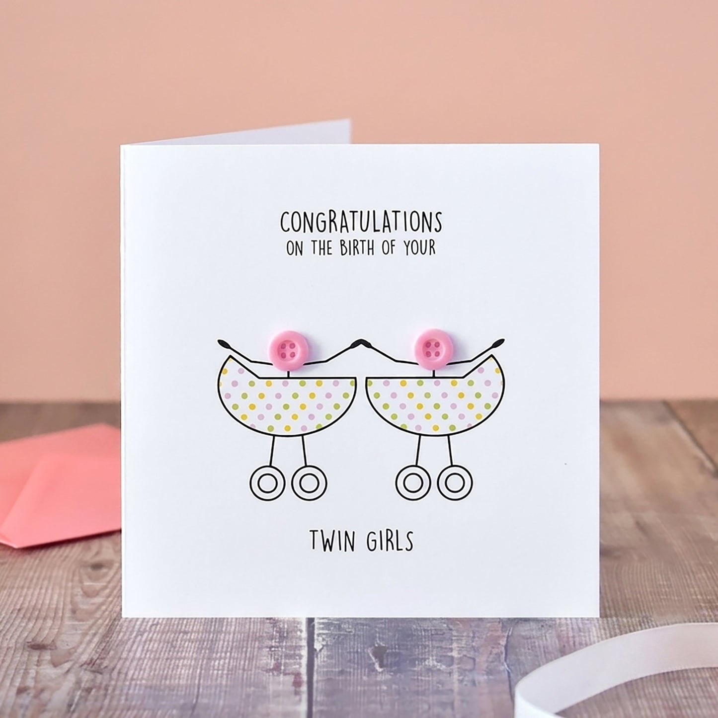 A white greeting card standing on a wooden surface, printed with the message 'Congratulations on the birth of your twin girls' and a simple line drawing of two baby prams with pink bows. A pink envelope sits behind it, with a vase of pale flowers and a pink ribbon in the background.