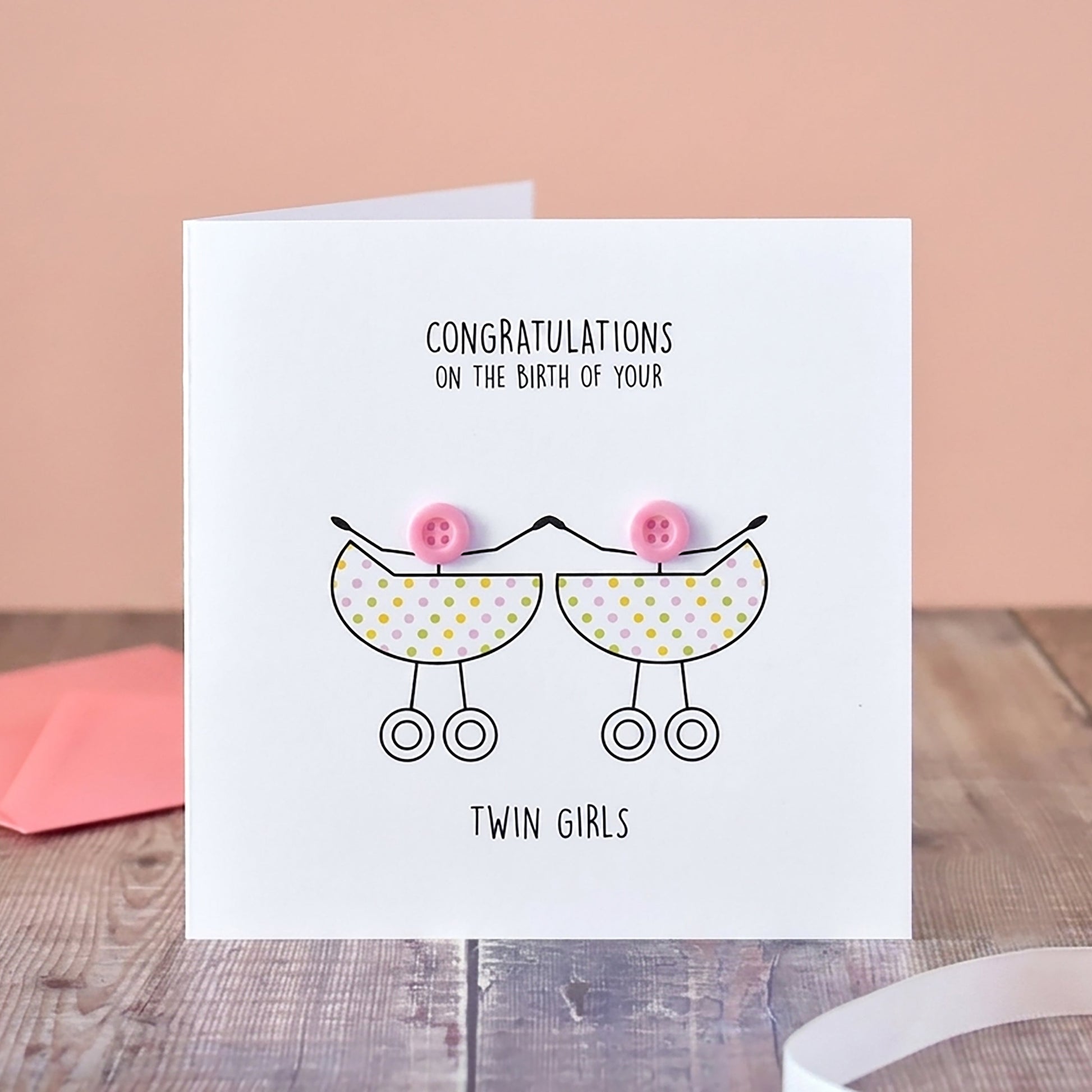 A white greeting card standing on a wooden surface, printed with the message 'Congratulations on the birth of your twin girls' and a simple line drawing of two baby prams with pink bows. A pink envelope sits behind it, with a vase of pale flowers and a pink ribbon in the background.