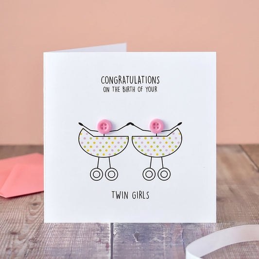 A white greeting card standing on a wooden surface, printed with the message 'Congratulations on the birth of your twin girls' and a simple line drawing of two baby prams with pink bows. A pink envelope sits behind it, with a vase of pale flowers and a pink ribbon in the background.