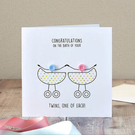 A square white congratulations card propped on a wooden surface, featuring a simple line drawing of two baby prams with colourful polka dots and the text 'CONGRATULATIONS ON THE BIRTH OF YOUR TWINS, ONE OF EACH!' A matching envelope lies to the right, with pink ribbon in the foreground against a beige wall background.