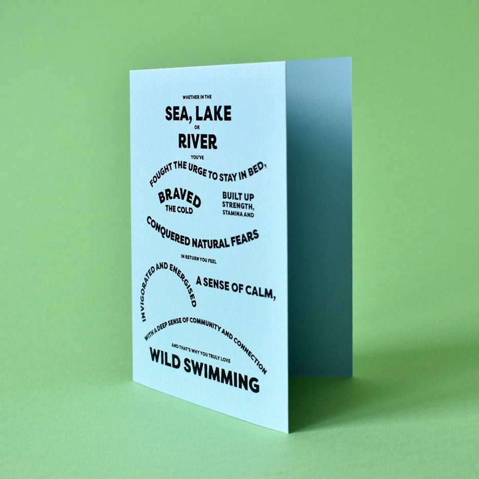 A folded greeting card standing upright on a solid light green background. The card is white with black text that reads 'WILD SWIMMING' and 'SEA, LAKE RIVER', surrounded by smaller black text and circular graphic elements.